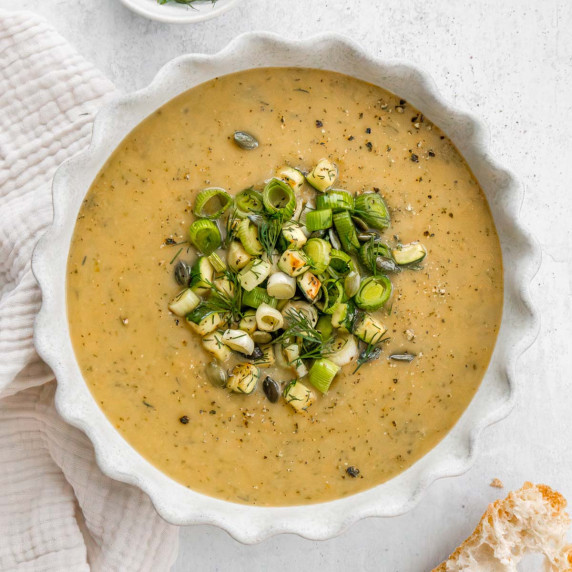 Zucchini and leek soup in a cream fluted bowl with a napkin and crusty bread. 